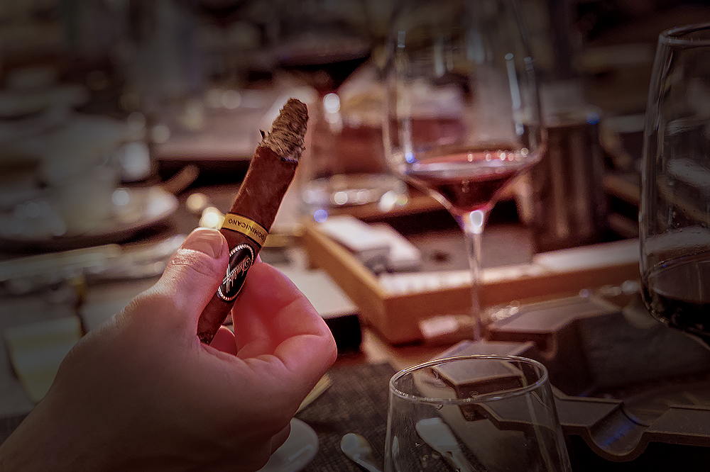 Konzentrierter Genuss in der Nahaufnahme: Die Davidoff Puro Dominicano brennt gleichmäßig ab und offenbart ihre weiße, stabile Asche. Im Hintergrund wartet der tiefrote Walliser Wein darauf, die Schokoladen- und Röstaromen der Zigarre auf der Zunge zu untermalen.